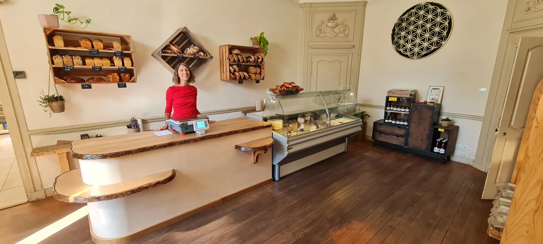 Une commerçante derrière le comptoir en bois de l'épicerie, avec les pains rangés en cagettes au mur et la vitrine des fromages à droite.